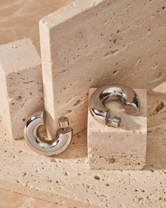  A pair of platinum Perfect Hoop Grassetto earrings is displayed on an assortment of stone slabs and blocks, the chunky open hoop of each earring gleaming as it reflects its surroundings. 