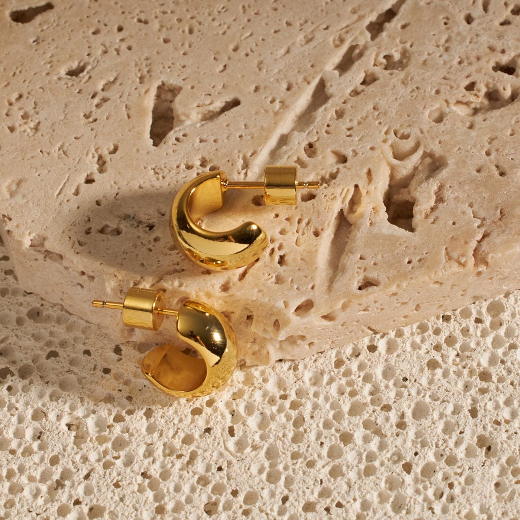 Small, yet bold, a pair of Nice Hoop earrings show off their wide C-curve design and pushback closure as they are displayed side-by-side on a stone slab. 
