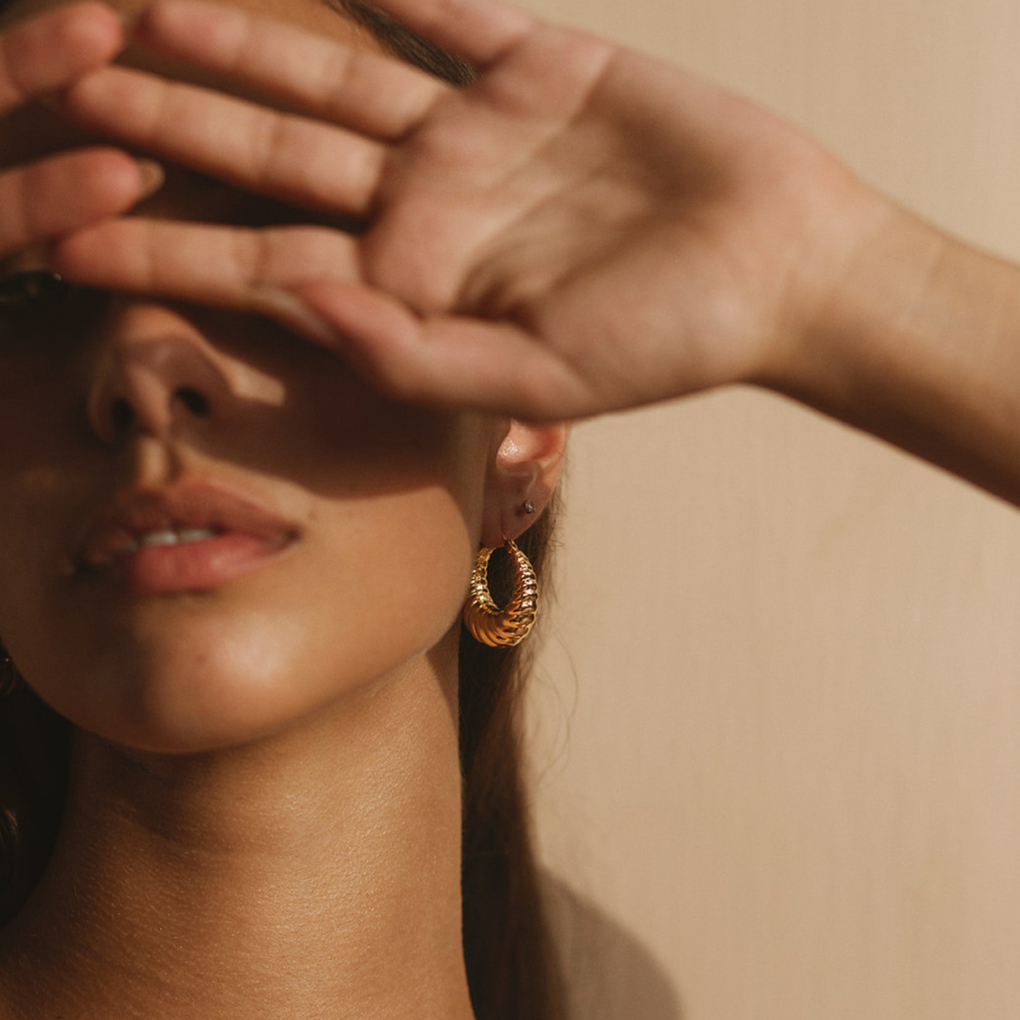 The model shades her eyes with her hand, drawing attention to the golden mid-sized hoop on her ear. 
