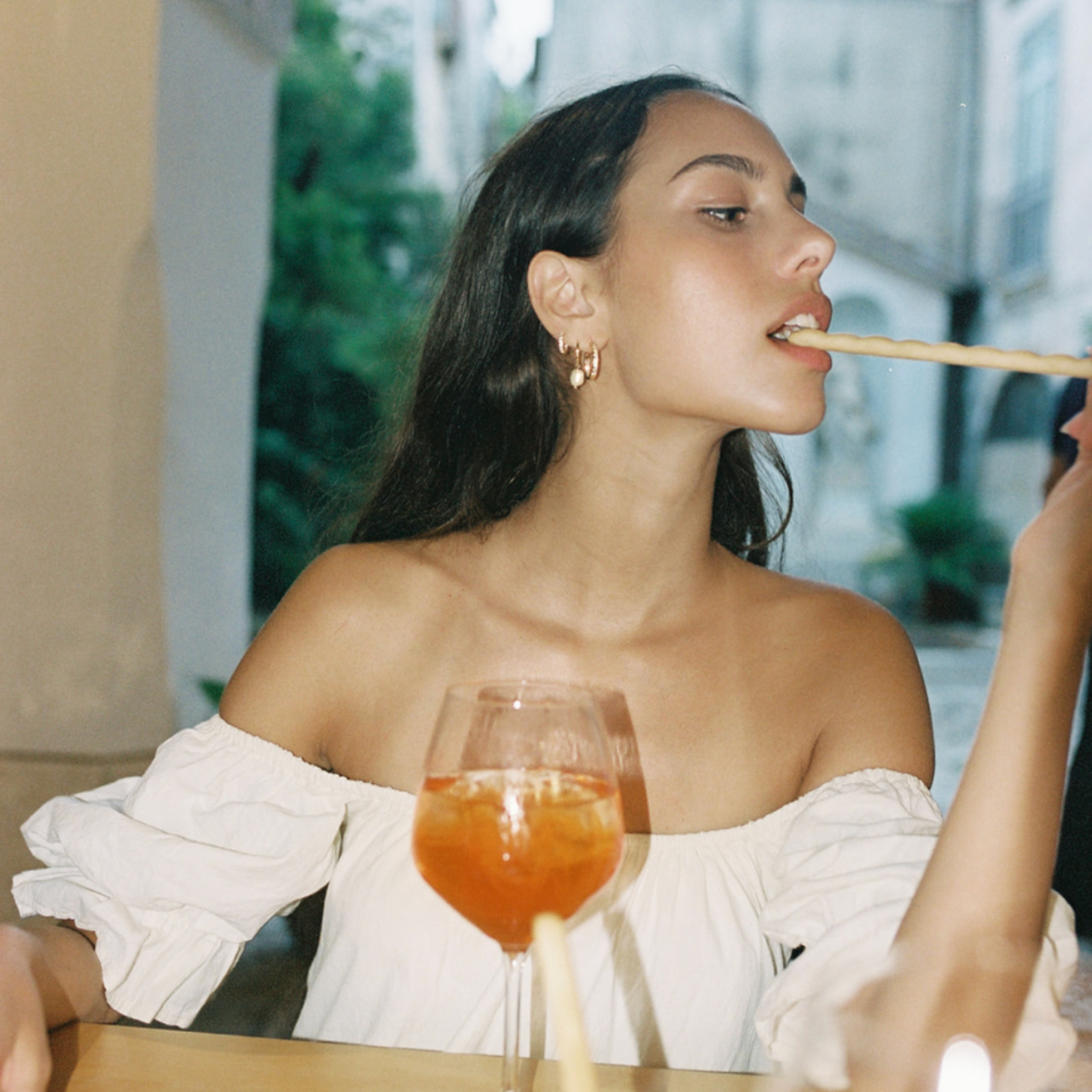 The model stretches to bite a rolled waffle snack, a cocktail before her, and a mini hoop and stud earring stack on her ear.