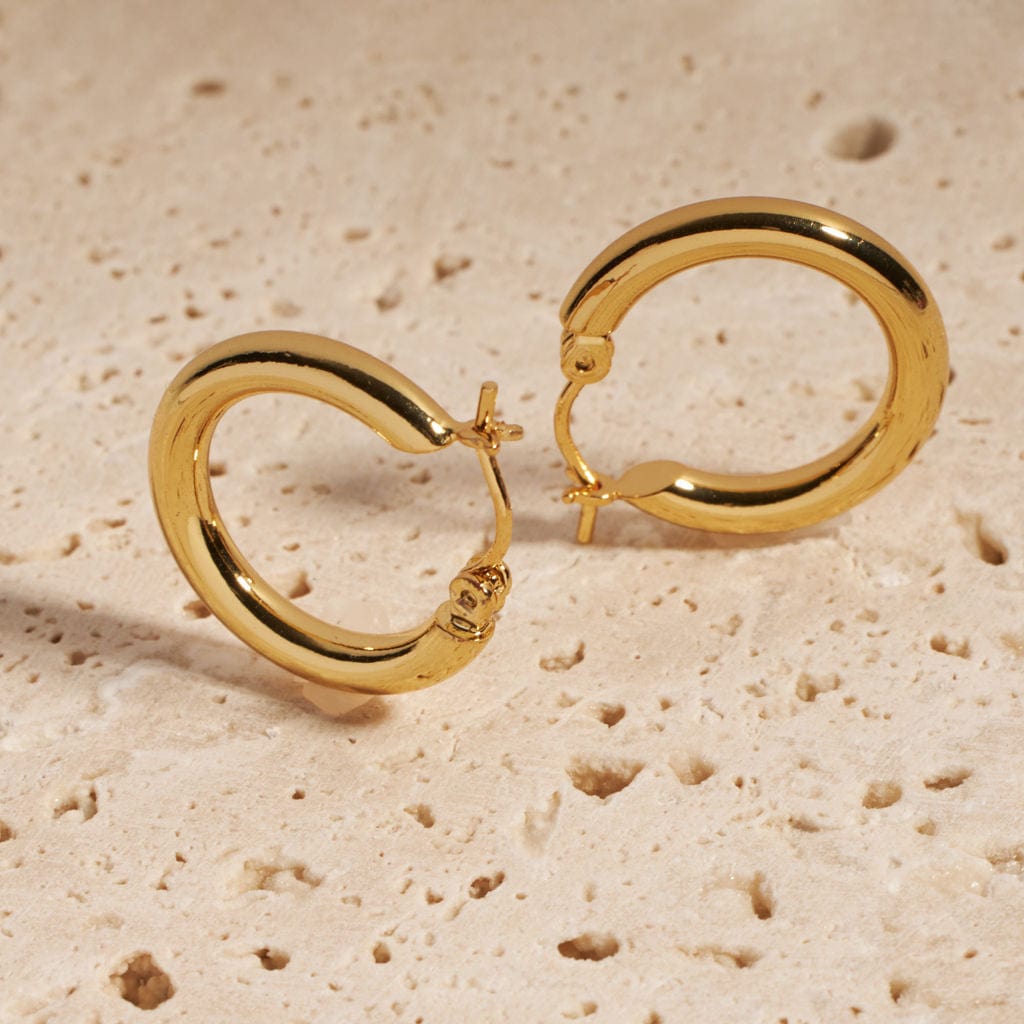 A pair of Ravello Hoop earrings stands together on a porous stone slab, each earring propped up on its perfectly round, golden curve.