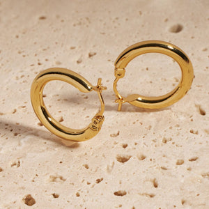 A pair of Ravello Hoop earrings stands together on a porous stone slab, each earring propped up on its perfectly round, golden curve.