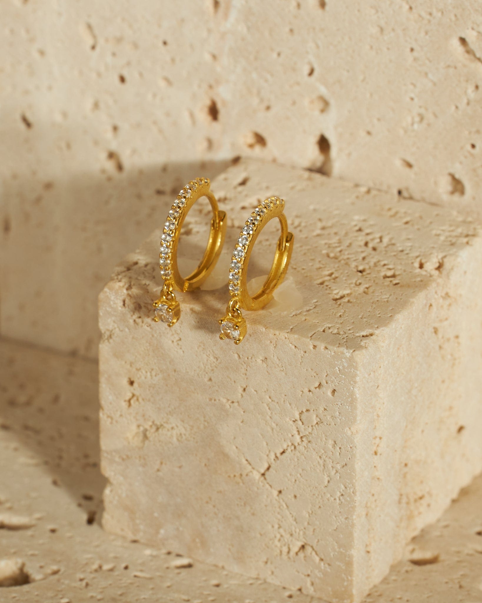 A pair of crystal-encrusted Bellagio Drop Hoop earrings sits atop a stone block, the larger crystal pendants dangling gracefully from the bottom of each hoop.