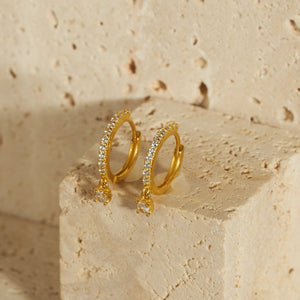 A pair of golden Bellagio Hoop earrings sit side by side on a porous stone block, the row of crystal accents sparkling where they catch the light as the larger crystal charms hang suspended along the side of the block. 