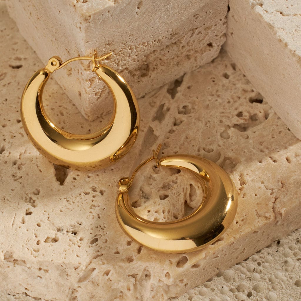 A pair of Capri Hoop earrings is displayed on a stone surface, the golden hoops heavy at their base while the tops narrow to a sleek latch back closure. 