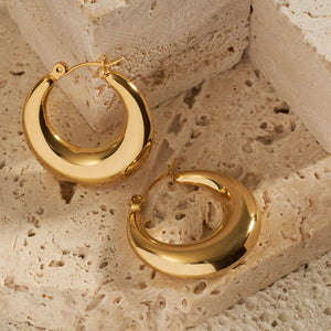 A pair of Capri Hoop earrings is displayed on a stone surface, the golden hoops heavy at their base while the tops narrow to a sleek latch back closure. 