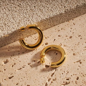 Two Perfect Hoop Midi earrings are displayed against a porous stone slab, the golden curve of each hoop shining where it catches the light.