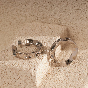Two platinum Perfect Hoop Grassetto earrings are displayed on a porous stone surface, the hammered texture catching both light and shadows around the shining silver curves. 