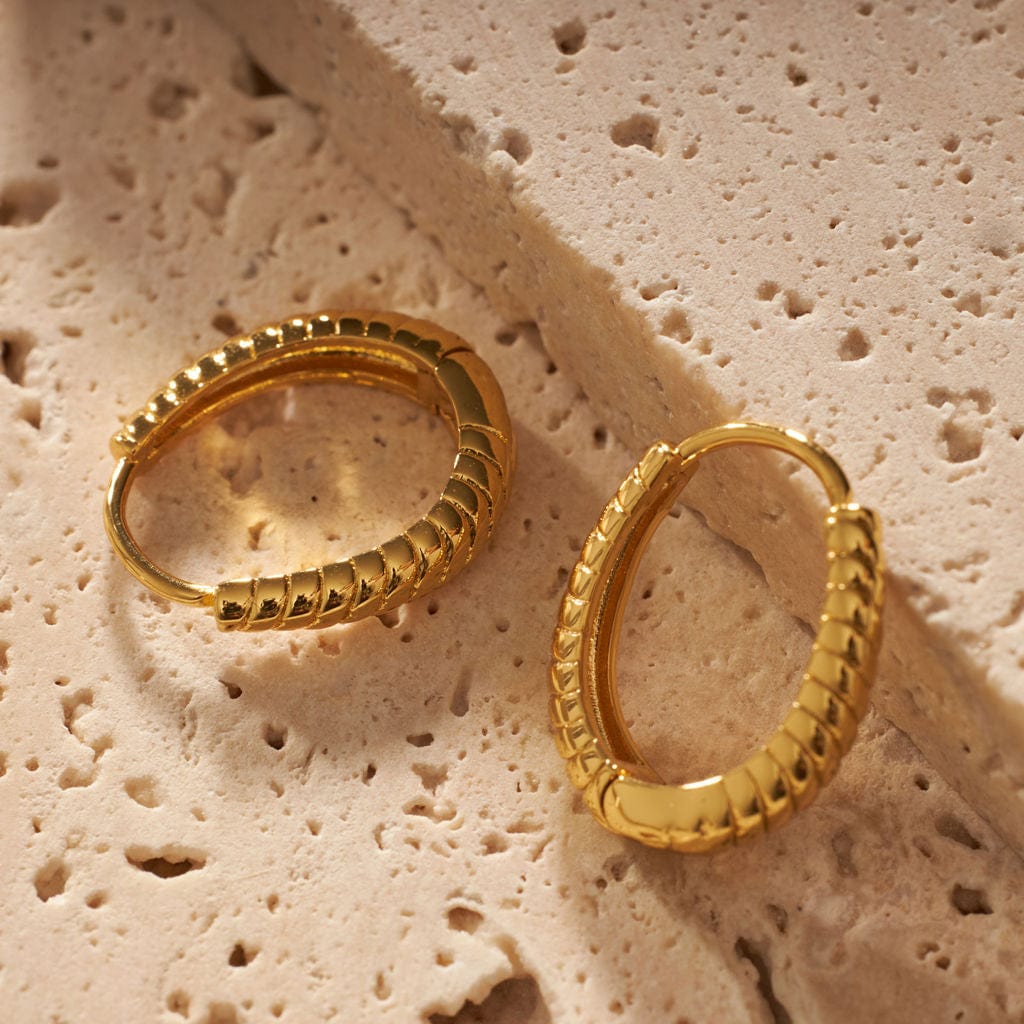 A pair of Saint Germain Hoop earrings show off their golden glow and segmented design as they lay atop a porous stone slab. 