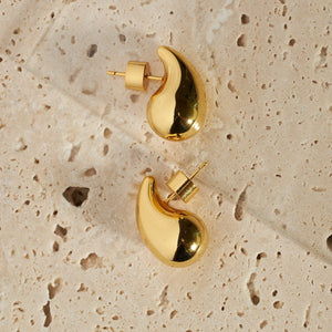 A pair of Moda Hoop Minis lay on a porous stone surface, the golden cylinder backings showing from behind the gleaming, teardrop-shaped open hoop earrings.