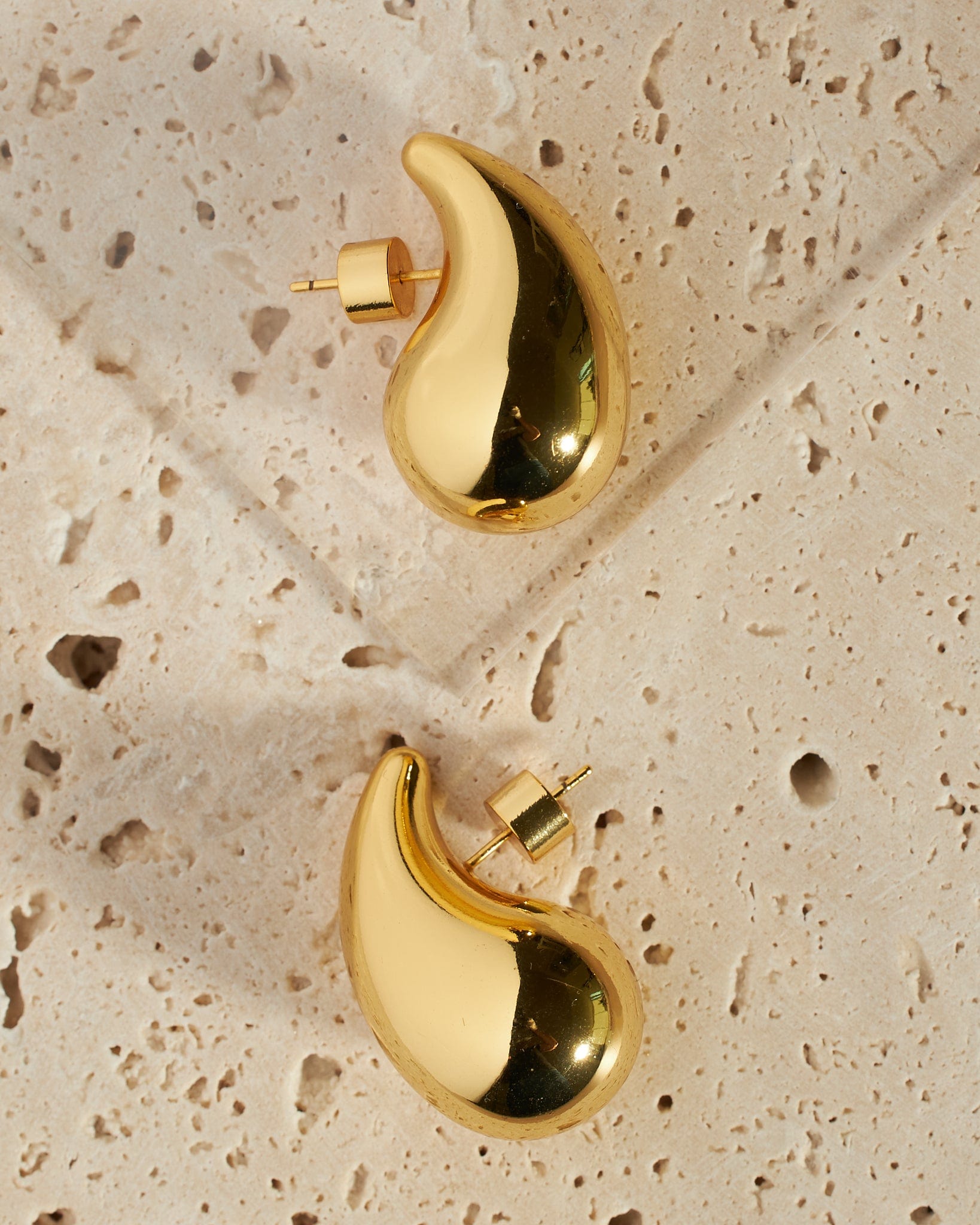 The teardrop shape and smooth, golden surface of a pair of Moda Hoop earrings stand out against the white stone slabs they rest on. 