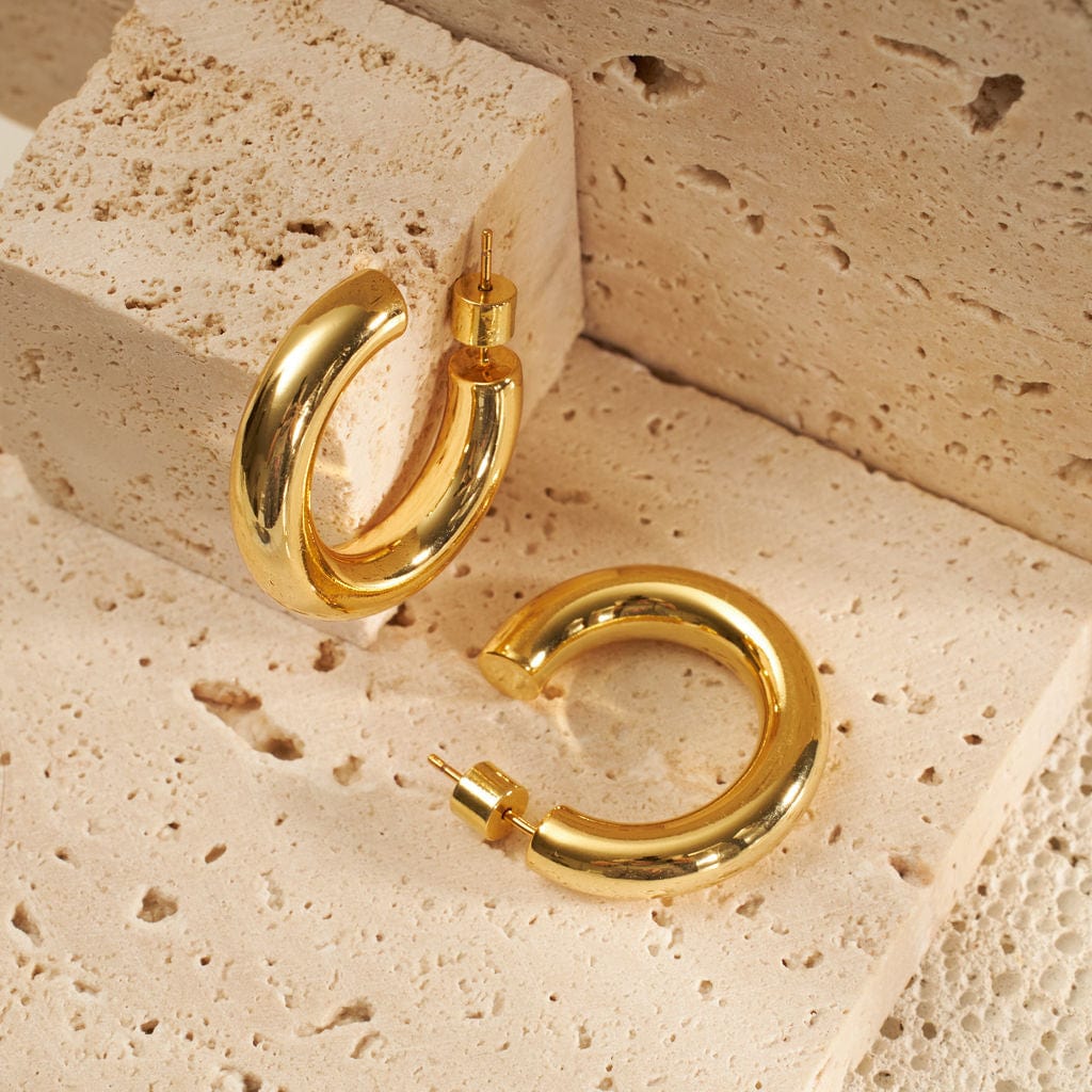 One golden Perfect Hoop earring is propped up on its curve against a stone block while another lays adjacently on its side. 