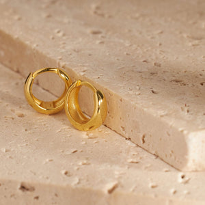 Two Capri Huggie earrings sit next to each other on a stone slab, each propped up on its wide curve to show the hollow interior and snug lever back closure. 