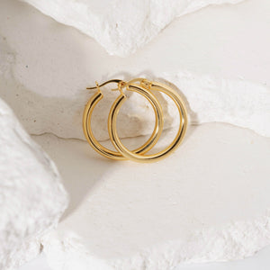 A pair of Allegra Hoop earrings sits together against a white stone slab, showcasing the golden curves and lever back closures. By The Perfect Hoop
