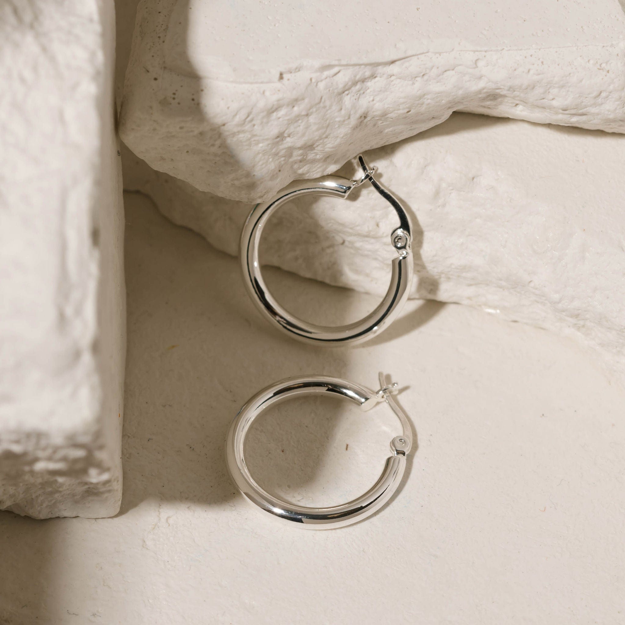 One sterling silver Allegra Hoop Midi lies on its side while the other stands against white stone slabs to showcase their perfectly circular shapes and lever back closures. 