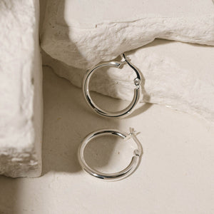 One sterling silver Allegra Hoop Midi lies on its side while the other stands against white stone slabs to showcase their perfectly circular shapes and lever back closures. 