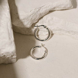 A pair of sterling silver Allegra Hoop Mini earrings shines in the light as they sit on some white stone slabs.  By The Perfect Hoop