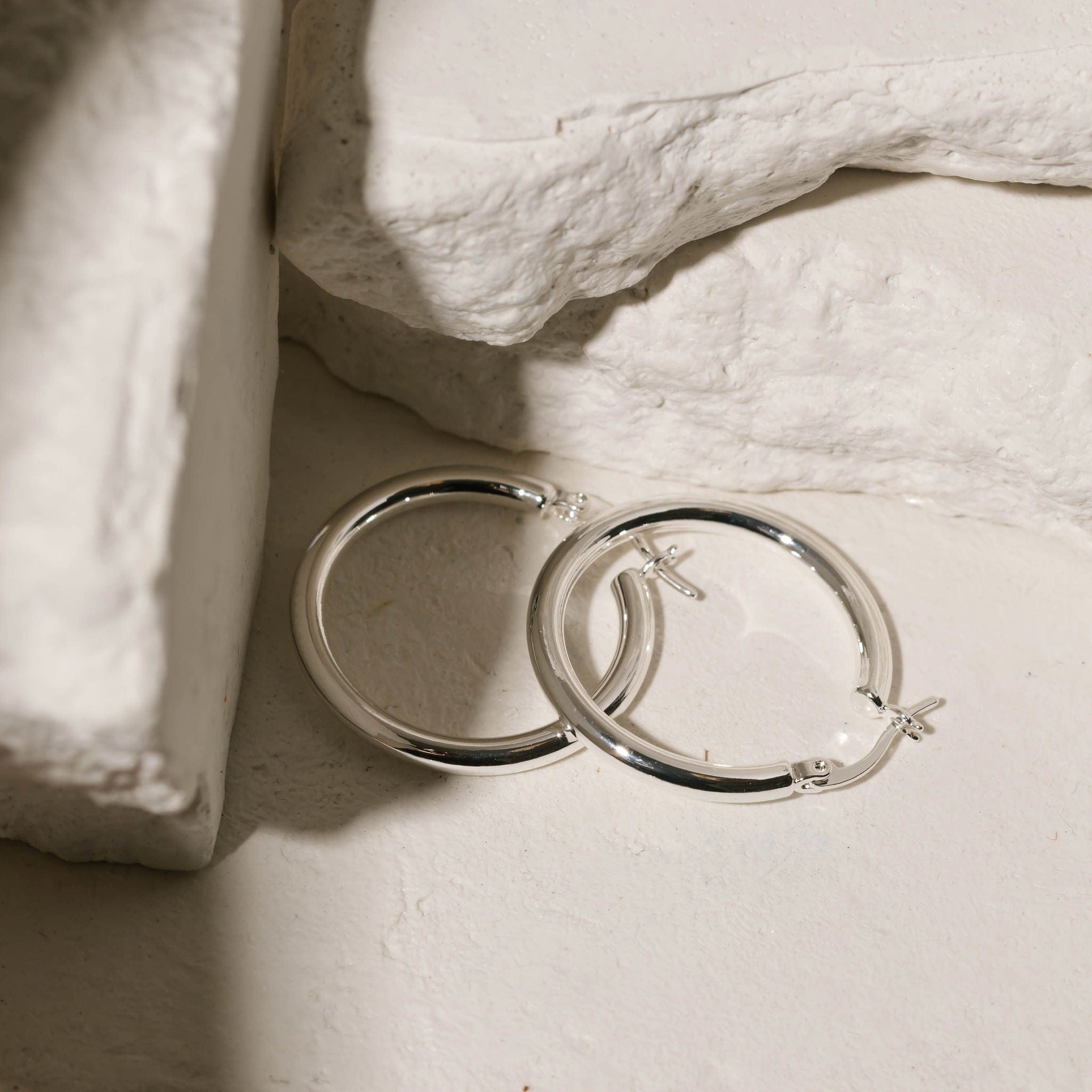 The perfectly circular shape and lever back closure of the sterling silver Allegra Hoop are clearly seen as two earrings lie together against white stone slabs. 