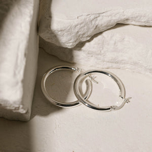 The perfectly circular shape and lever back closure of the sterling silver Allegra Hoop are clearly seen as two earrings lie together against white stone slabs. 