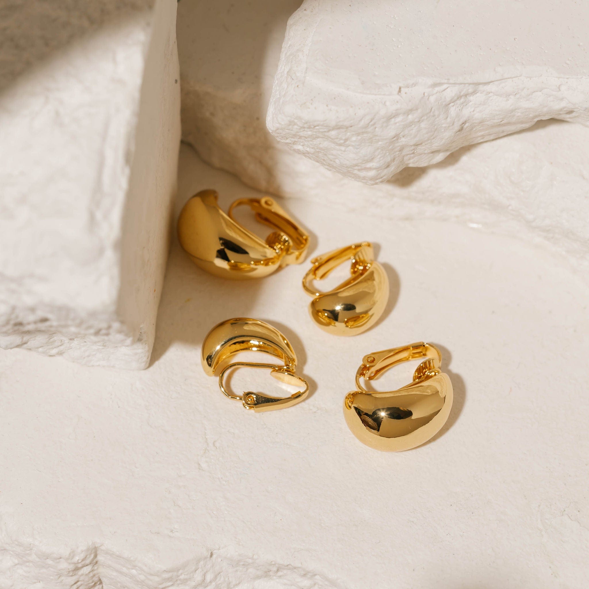 Gold Amadora Hoop Duo Clip-On earrings featuring a smooth, polished finish and a bold, curved design, displayed on a textured stone background. Elegant and versatile, these clip-on hoops by The Perfect Hoop offer a classic yet modern style