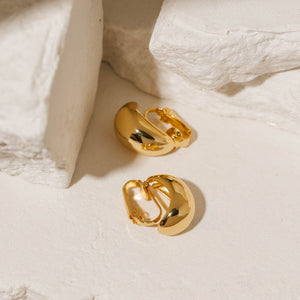 Gold Amadora Hoop Clip-On earrings featuring a smooth, polished finish and a classic, bold design, elegantly arranged on a textured stone surface. These clip-on hoops by The Perfect Hoop provide a timeless and versatile accessory option