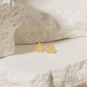 Gold heart-shaped stud earrings with crystal accents placed on a textured stone background, perfect for adding a touch of elegance to any look. Sparkling heart studs by The Perfect Hoop.