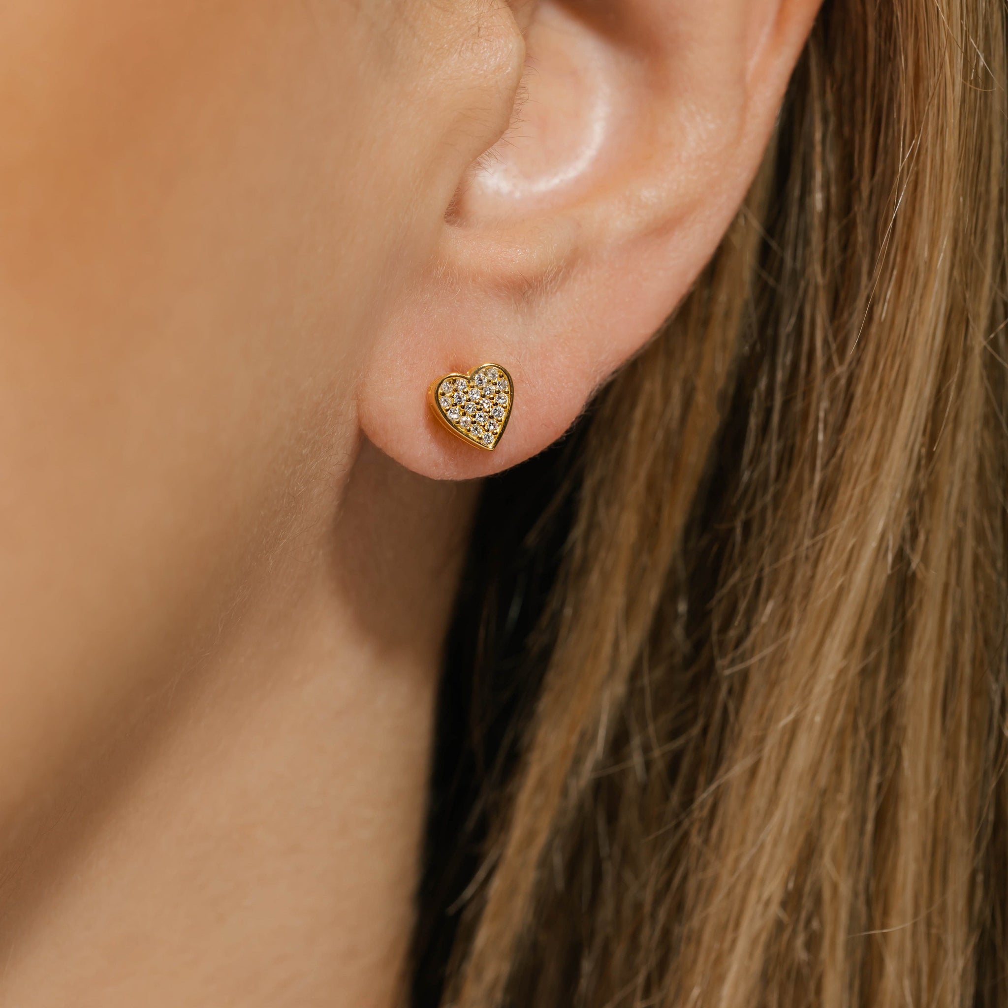 Model wearing gold pavé heart-shaped stud earrings with sparkling crystal accents, offering an elegant and refined look. Pavé heart stud earrings by The Perfect Hoop.