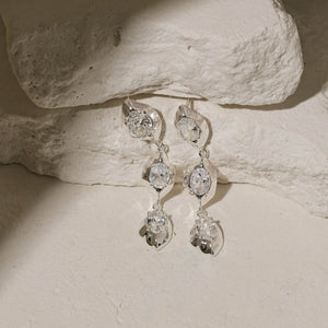 The crystal accents and sterling silver settings of a pair of Aura Hoop earrings shine brightly as the earrings lay against tiered stone slabs. 