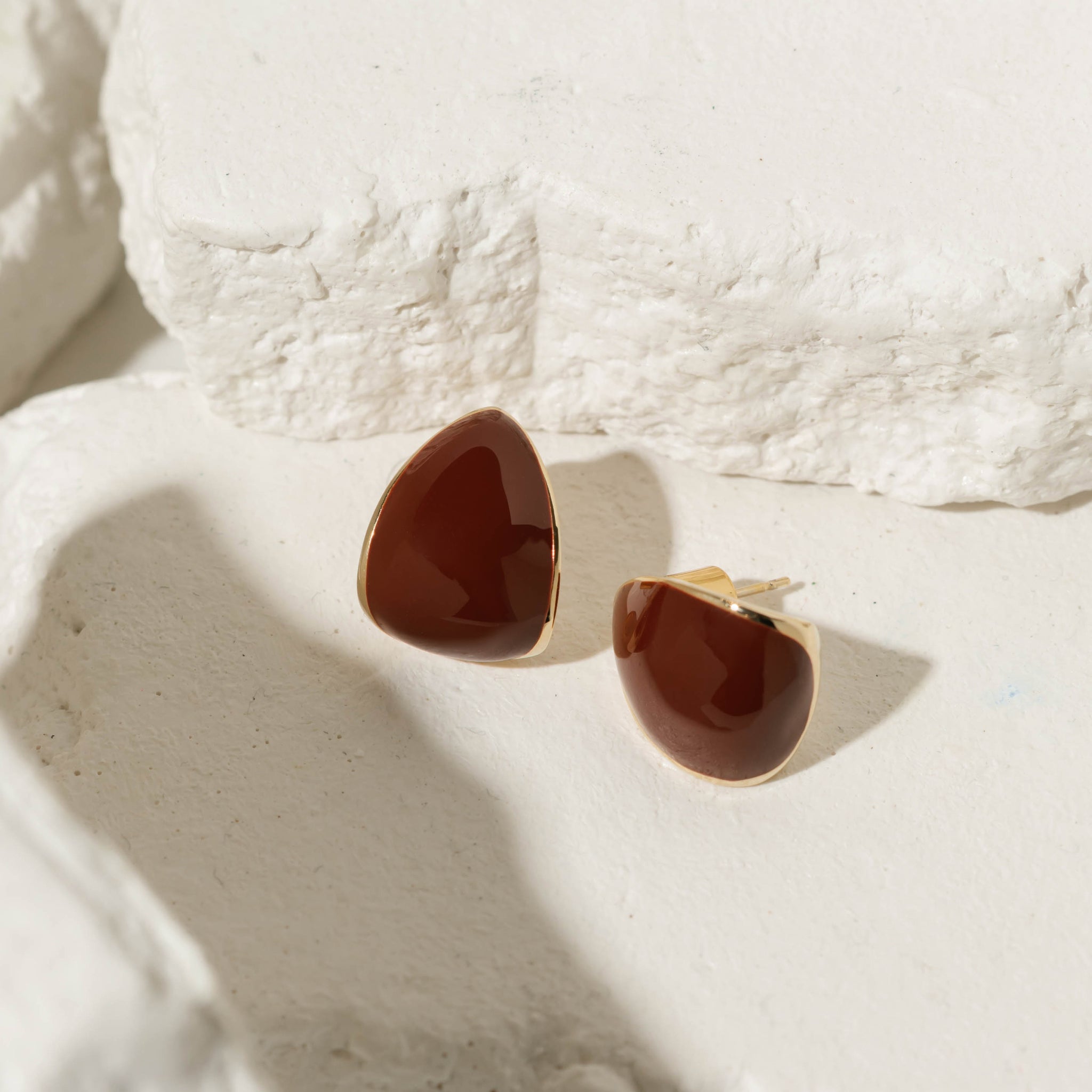 The Cabernet Hoop earrings featuring a deep red enamel finish with a soft triangular shape framed in gold, displayed on a textured stone surface for a bold and elegant style