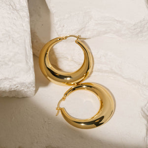 Gold Capri Hoop Grande earrings with a bold, rounded design and a smooth, polished finish, arranged on a textured stone surface. These statement hoop earrings by The Perfect Hoop bring a touch of glamour and sophistication.