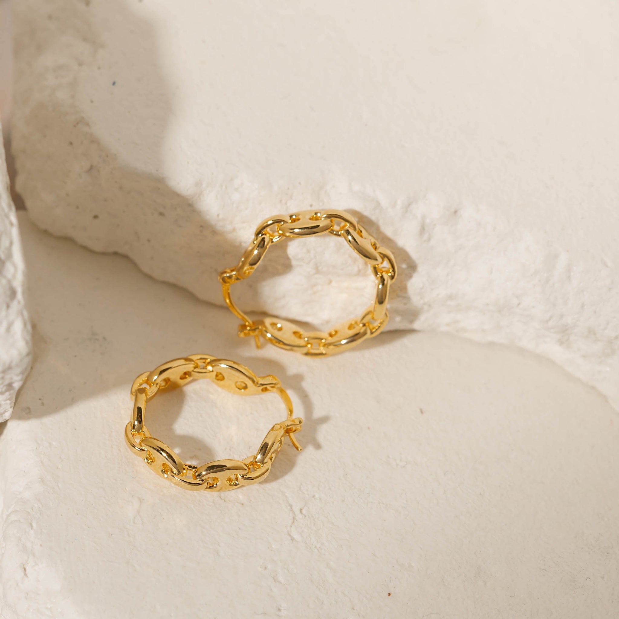 A golden Cavalla Hoop lay on its side as its pair stands balanced against a stone slab, each showcasing its chain link design and latch back closure. 