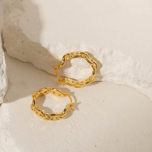 A golden Cavalla Hoop lay on its side as its pair stands balanced against a stone slab, each showcasing its chain link design and latch back closure. 