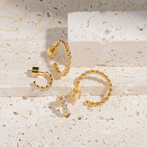 The Coquille Hoop Duo is displayed together against several stone blocks. One of each earring stands tall to display the sparkling crystals and gold settings, while the other two earrings show off the open hoop shape and push-back closures as they lay on their sides. 