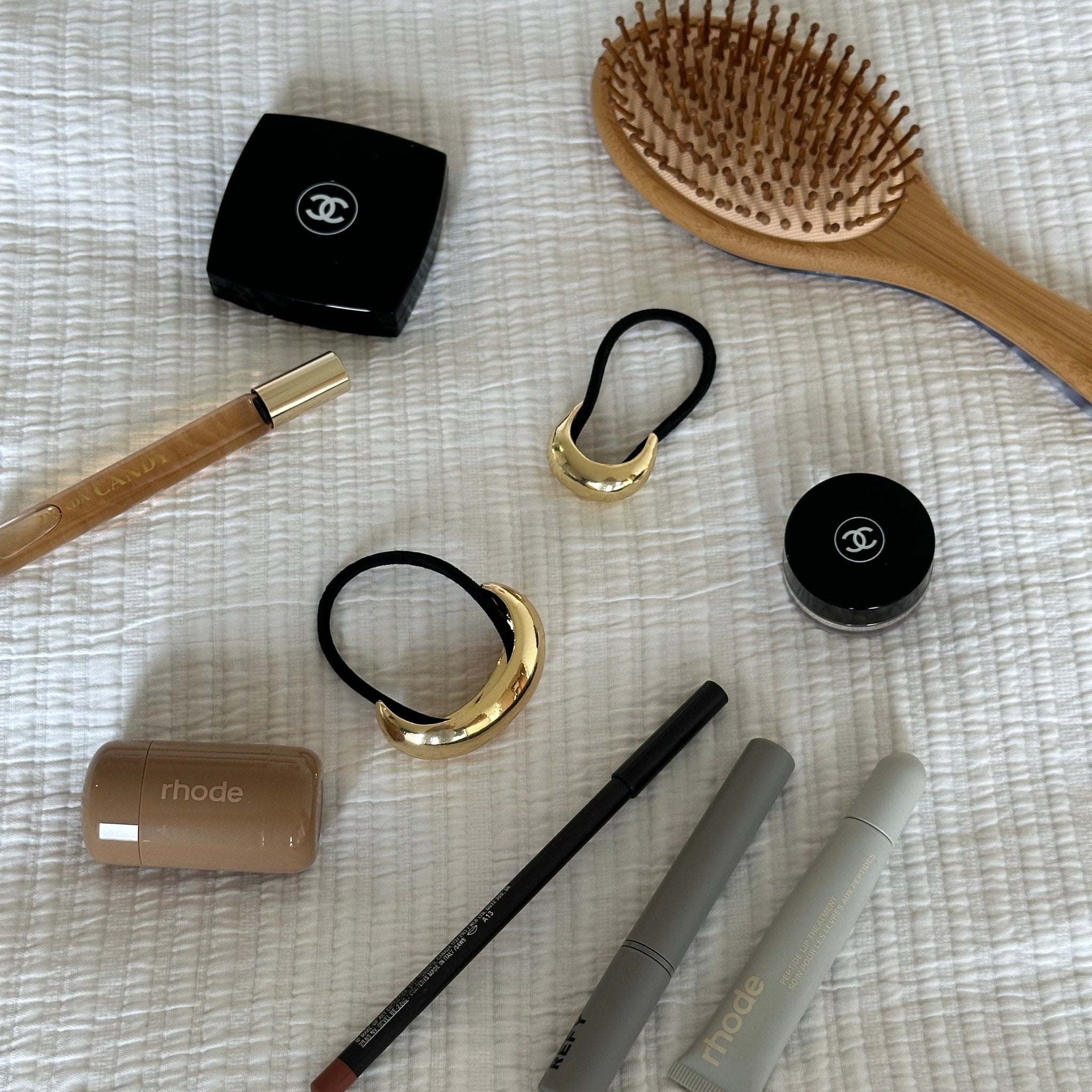 The Perfect Hair Hoop in polished gold shown as a duo on a textured white fabric, styled with luxury beauty essentials including Rhode, Chanel, and Caudalie, alongside a wooden paddle brush.