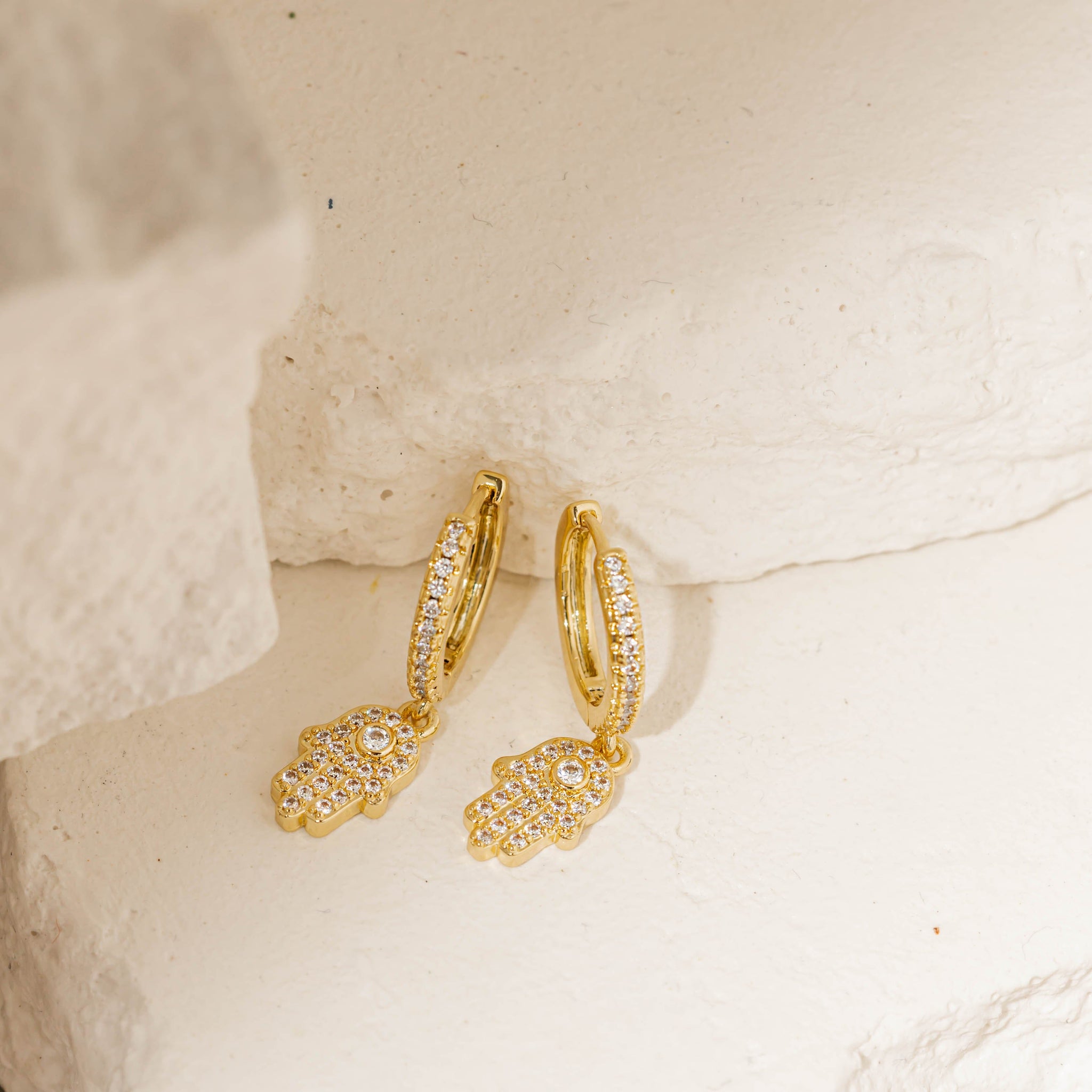 Gold pavé hoop earrings featuring dangling hamsa hand charms encrusted with crystals, elegantly displayed on a textured stone surface. Pavé hamsa hoop earrings by The Perfect Hoop.