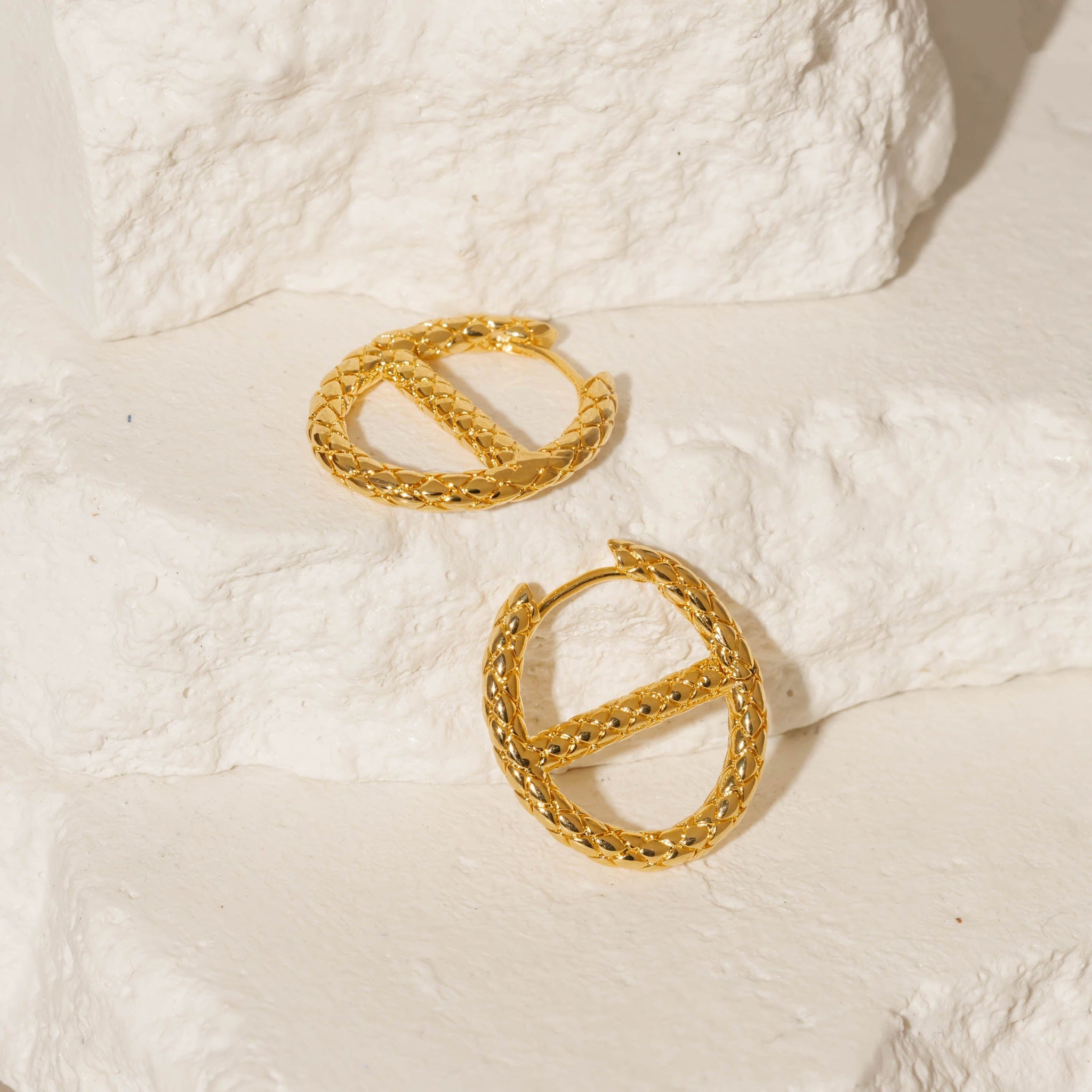 A pair of Jolie Hoop earrings lay side by side against white stone slabs, the crosshatch pattern exemplified as each golden hoop glistens in the light. 