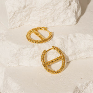 A pair of Jolie Hoop earrings lay side by side against white stone slabs, the crosshatch pattern exemplified as each golden hoop glistens in the light. 