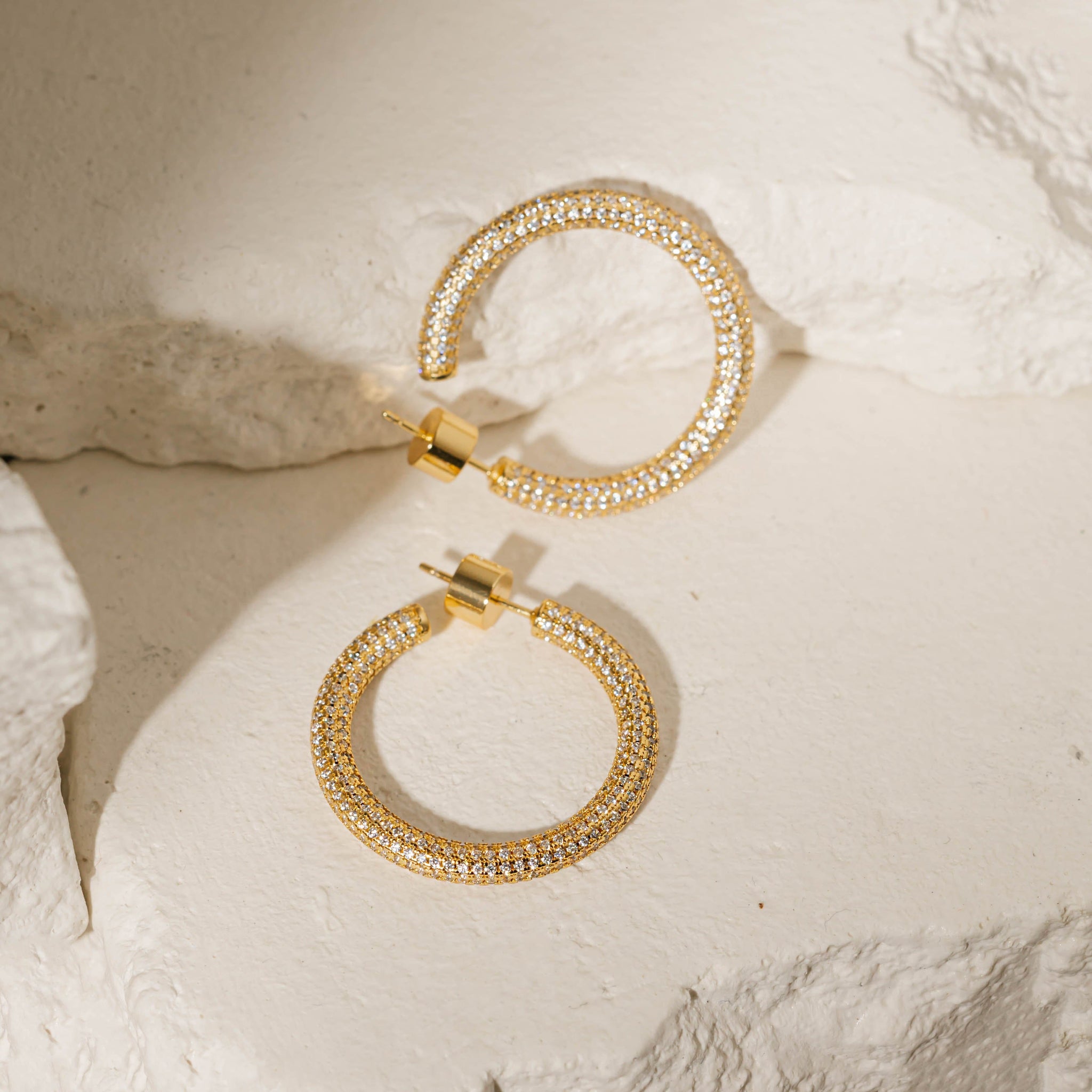 A pair of Malta Hoop Pavé earrings lay against a stone slab to display the open hoop design, cylindrical push back closures, and crystal accents that sparkle brightly in the light. 