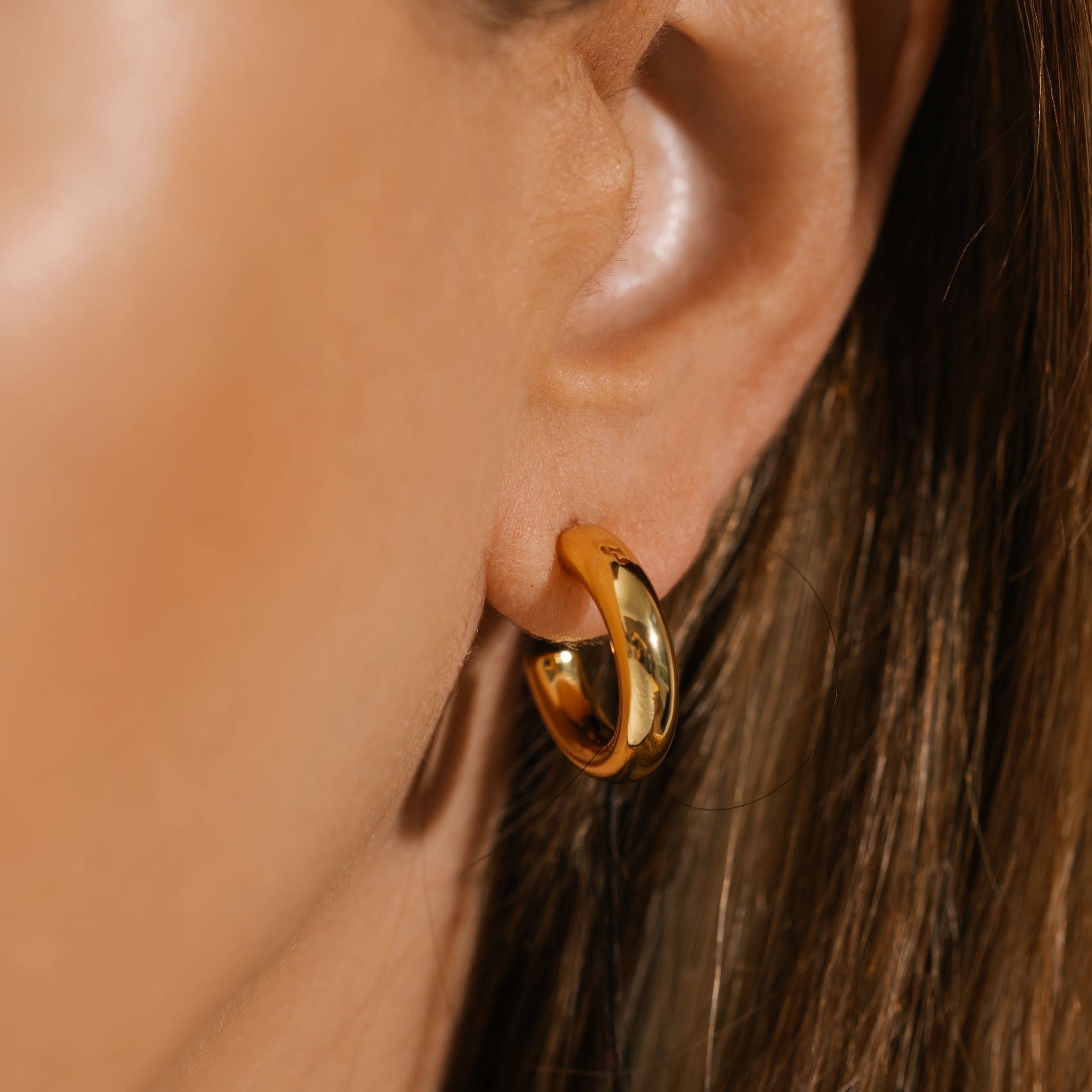 A close-up view of the Malta Hoop 20mm on the model's ear shows the open hoop design and the gleaming, golden surface reflecting the surroundings.  By The Perfect Hoop.
