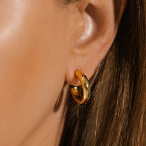 A close-up view of the Malta Hoop 20mm on the model's ear shows the open hoop design and the gleaming, golden surface reflecting the surroundings.  By The Perfect Hoop.