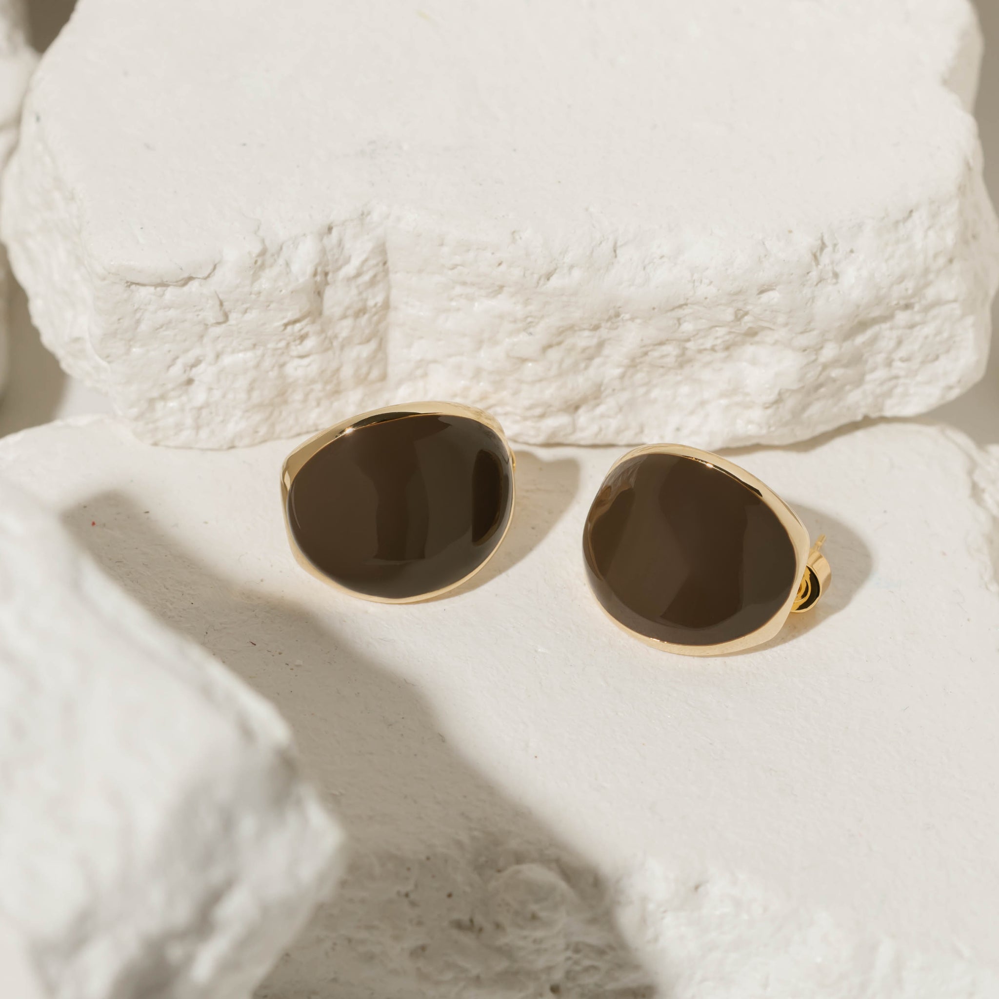 The Mocha Hoop earrings featuring a glossy chocolate brown enamel finish framed in gold, displayed on a textured stone surface for a refined and modern style.