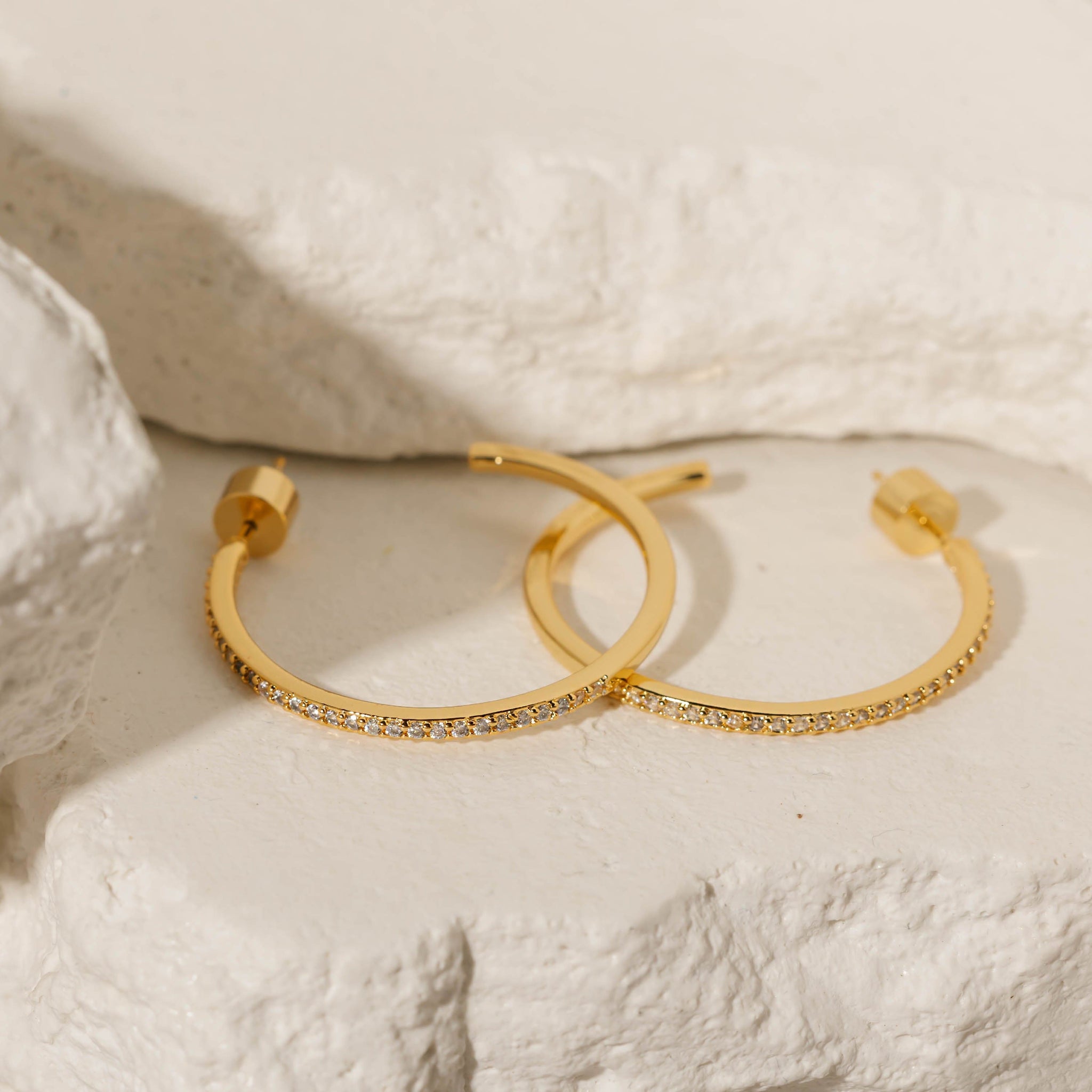 A pair of Monte Carlo Hoop earrings lay stacked together on a stone slab to show off the golden open hoops and sparkling crystal accents. 