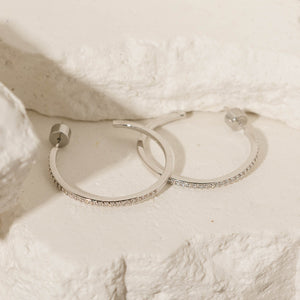 A pair of sterling silver Monte Carlo Hoop earrings lay together on a stone slab, displaying the open hoop design, crystal accents, and cylindrical pushback closures. 