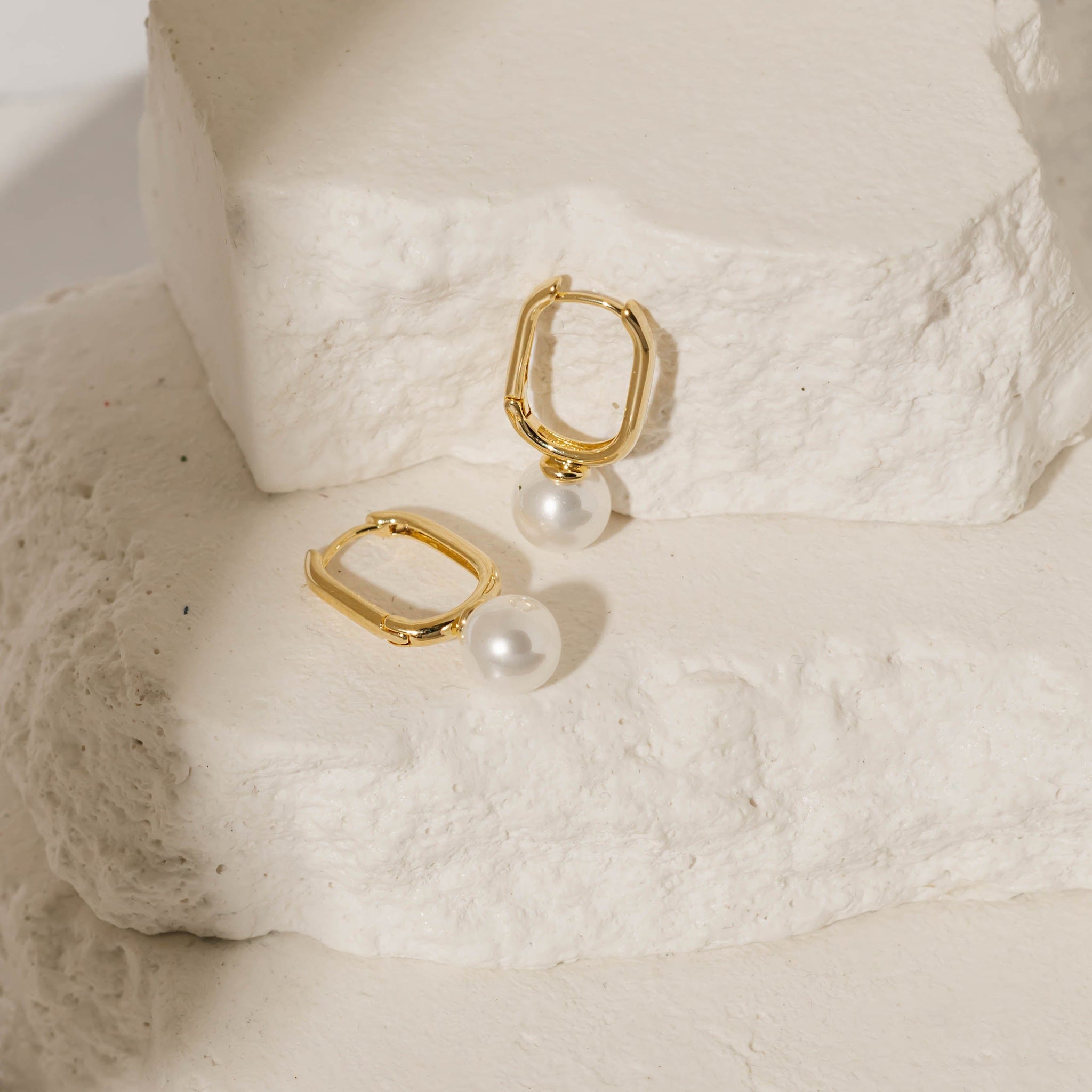 A pair of Ostra Hoop earrings is displayed against white stone slabs, one balancing on the pearl accent as the other lay flat on its side. 