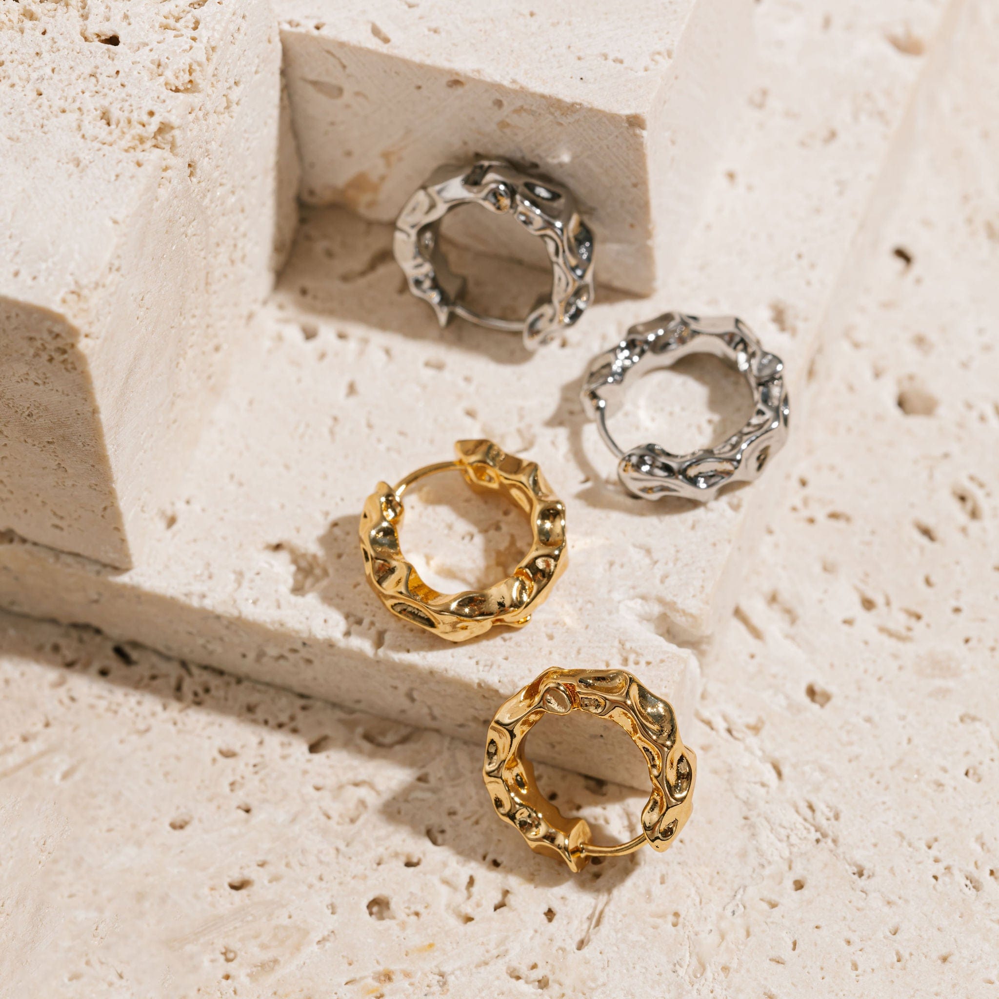  A pair of Marquis Hoop Mini earrings lies on an elevated counter while a pair of Marquis Hoop Mini Platinum earrings sits on the counter below, the hammered texture of each being accentuated in the light. 
