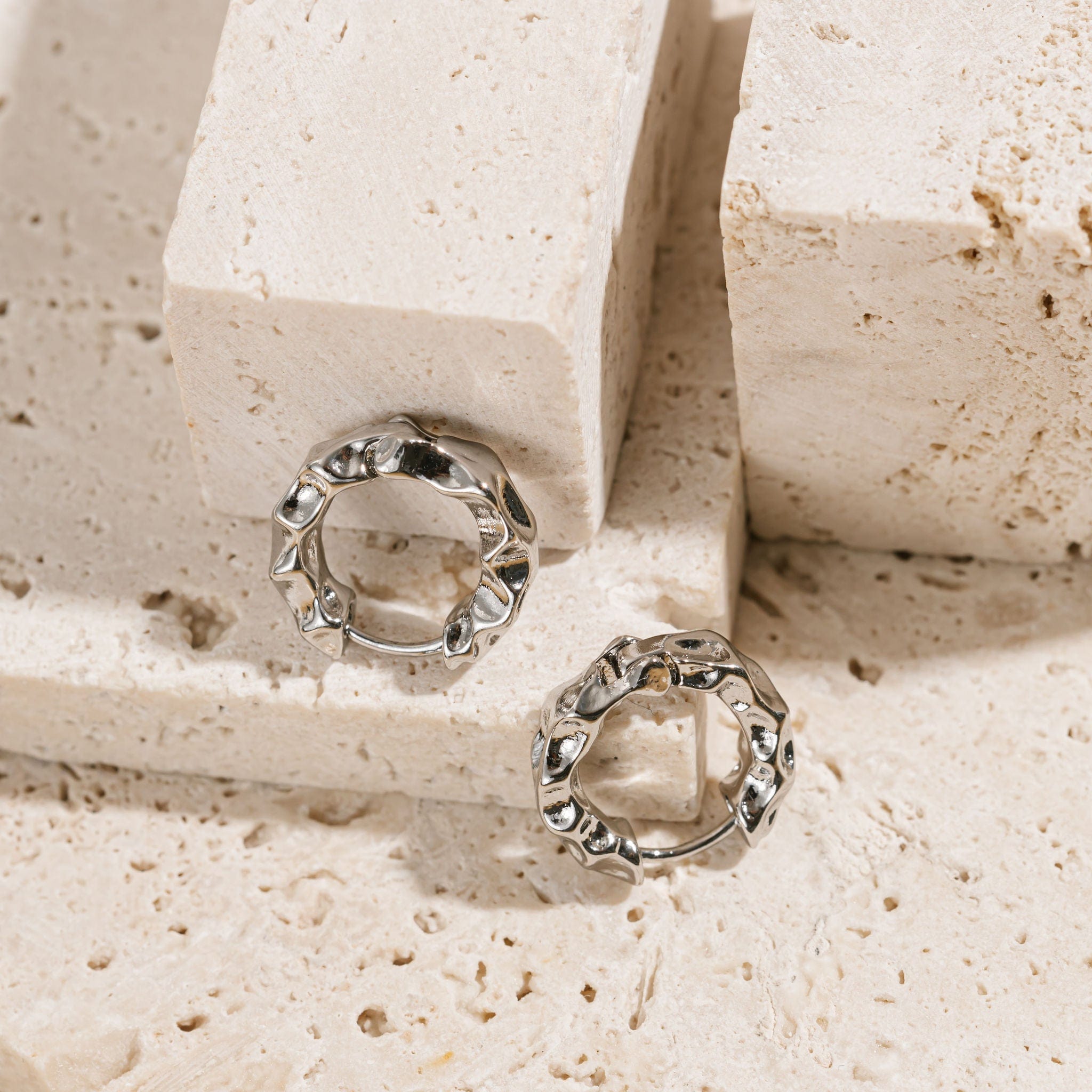  A pair of Marquis Hoop Mini Platinum earrings are displayed on a stone counter, the light accentuating the hammered texture of the hoops. 