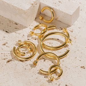 All four open hoop earrings in the Malta Hoop Quattro set lay on a porous stone slab, the various sizes laying within and atop one another while the smooth golden surfaces glisten in the light. 