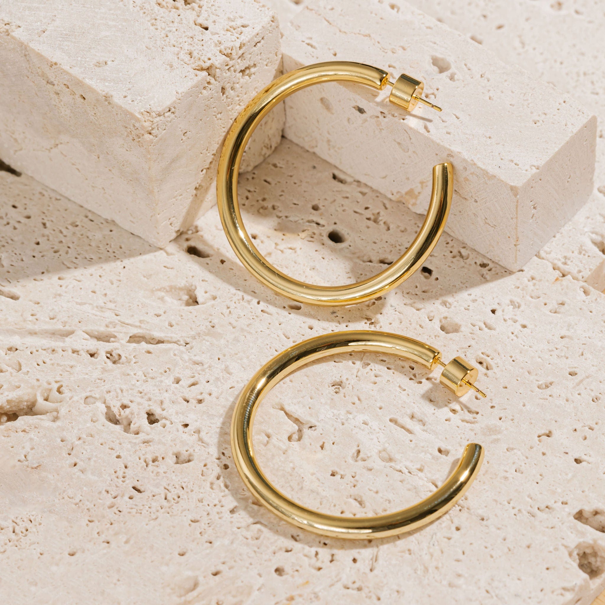 A pair of Malta Hoop 45mm earrings lays side by side on a porous stone slab, one earring laying flat on its side while the other is propped up against two raised blocks. 