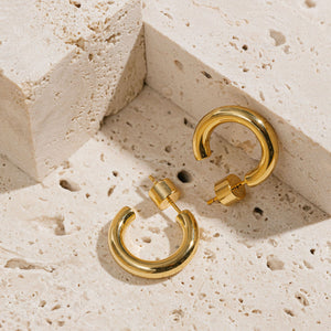 A pair of Malta Hoop 20mm earrings is displayed on a porous stone surface, one earring laying on its side while the other stands next to it on the cylindrical closure, propped against an elevated stone block.  