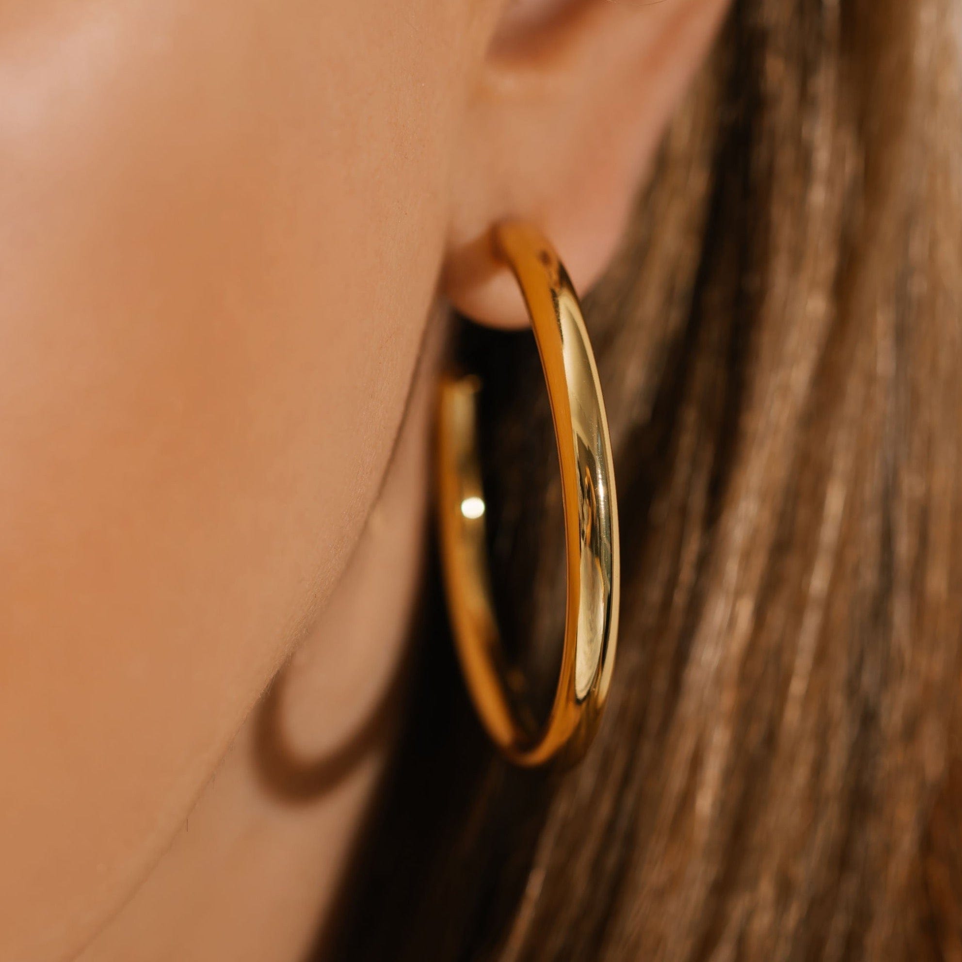 A close-up view shows the smooth and reflective golden surface of the large 45mm Malta Hoop earring as it's worn on the model's ear. 